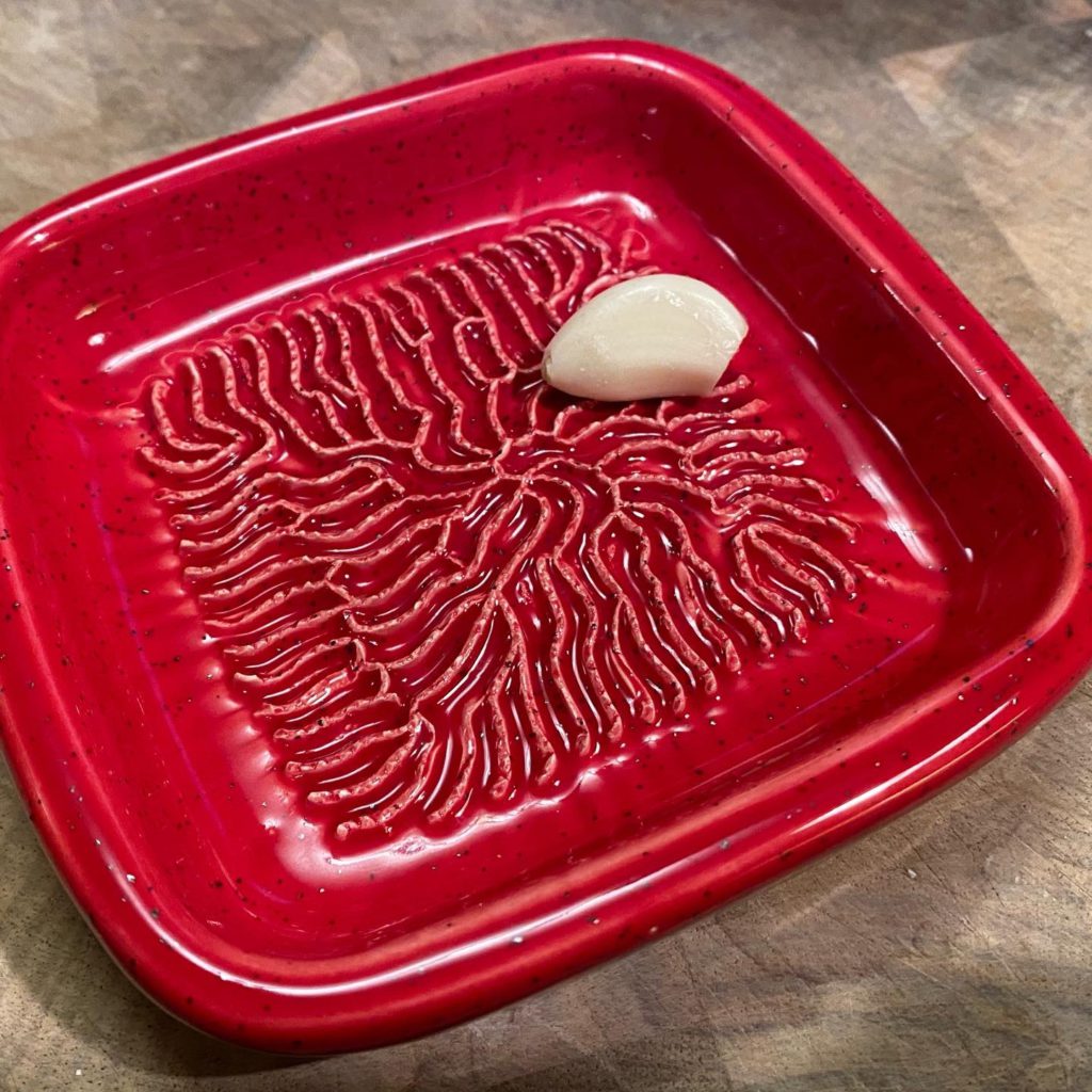 Red squarish ceramic dish with raised grooves on the bottom, with a clove of garlic