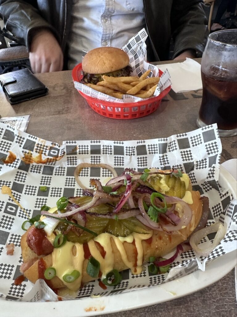 A hotdog covered in cheese and pickles, and a platter with a hamburger and fries