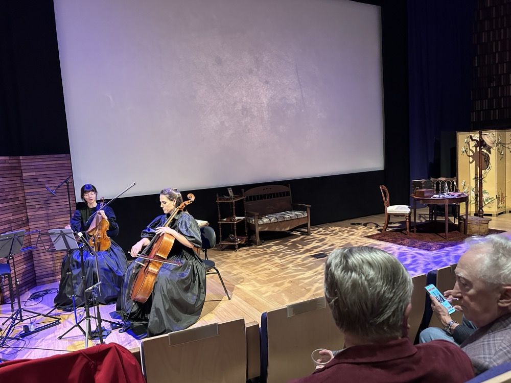 The stage of an auditorium. To the left is a string quartet of four women all dressed in black. To the right is a theatrical set with a table and chairs.