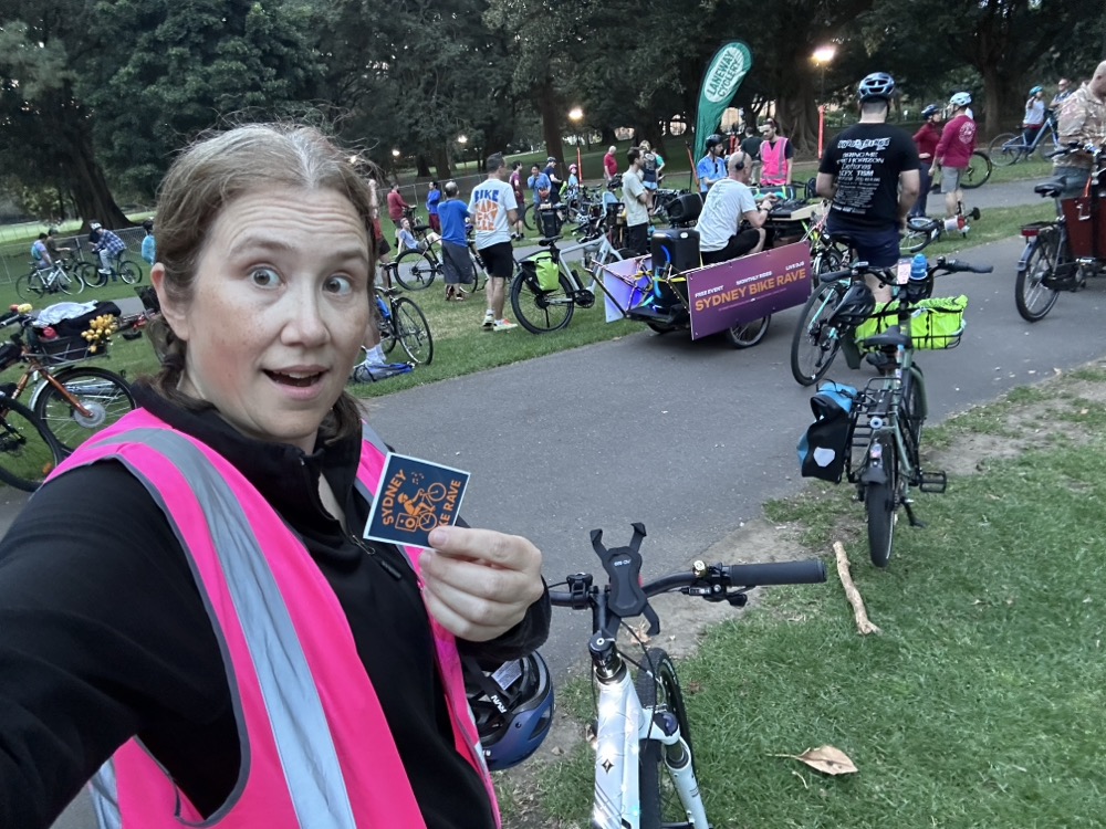 A cyclist in a pink reflective vest standing in a park. There's a bike trailer with a DJ on it.