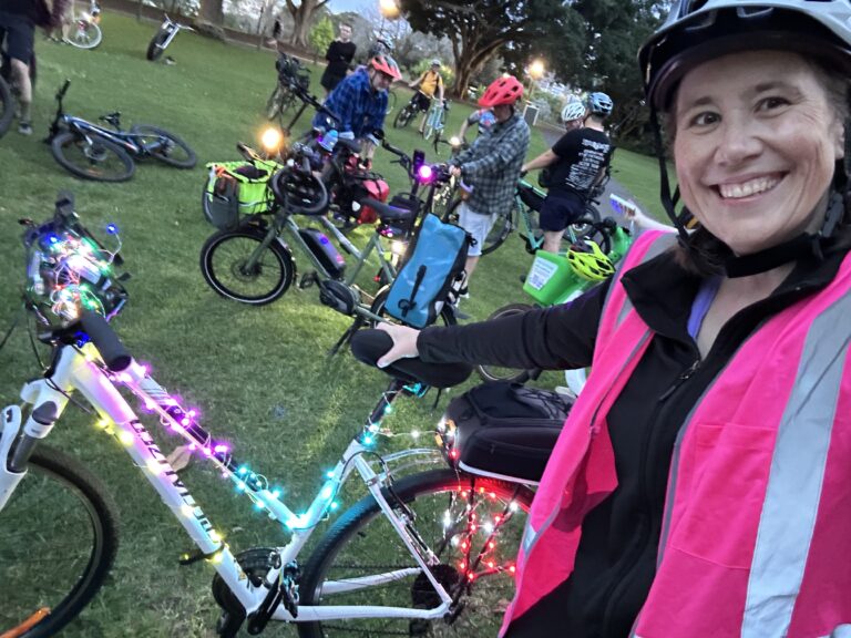 A woman cyclist in a pink reflective vest next to a bicycle covered in fairy lights