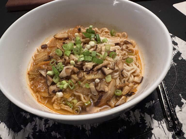 A large white bowl of kimchi ramen: ramen noodles with reddish broth, kimchi, shiitake mushrooms, and green onions