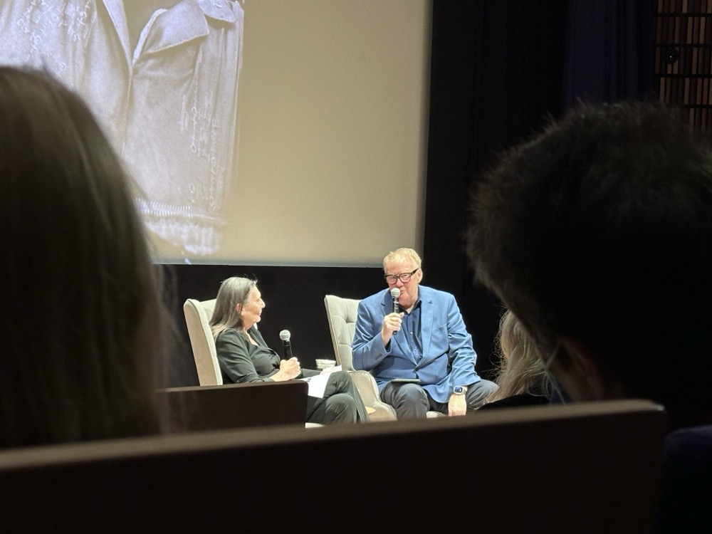 A man and a woman seated in chairs with microphones in front of an audience
