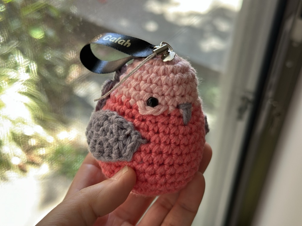 A small round pink crocheted bird with a ribbon attached to its head that says GALAH