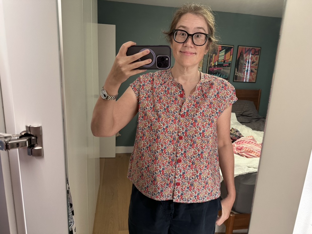 A mirror selfie of a woman in navy shorts and a sleeveless blouse in an allover print of tiny red, blue, and yellow flowers