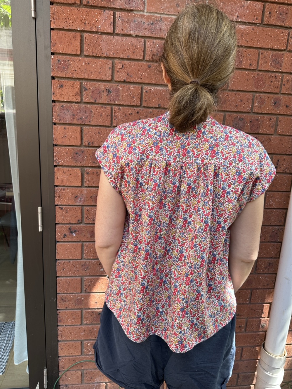 A rear view of a woman in navy shorts and a sleeveless blouse in an allover print of tiny red, blue, and yellow flowers. The blouse is gathered across the back yoke.