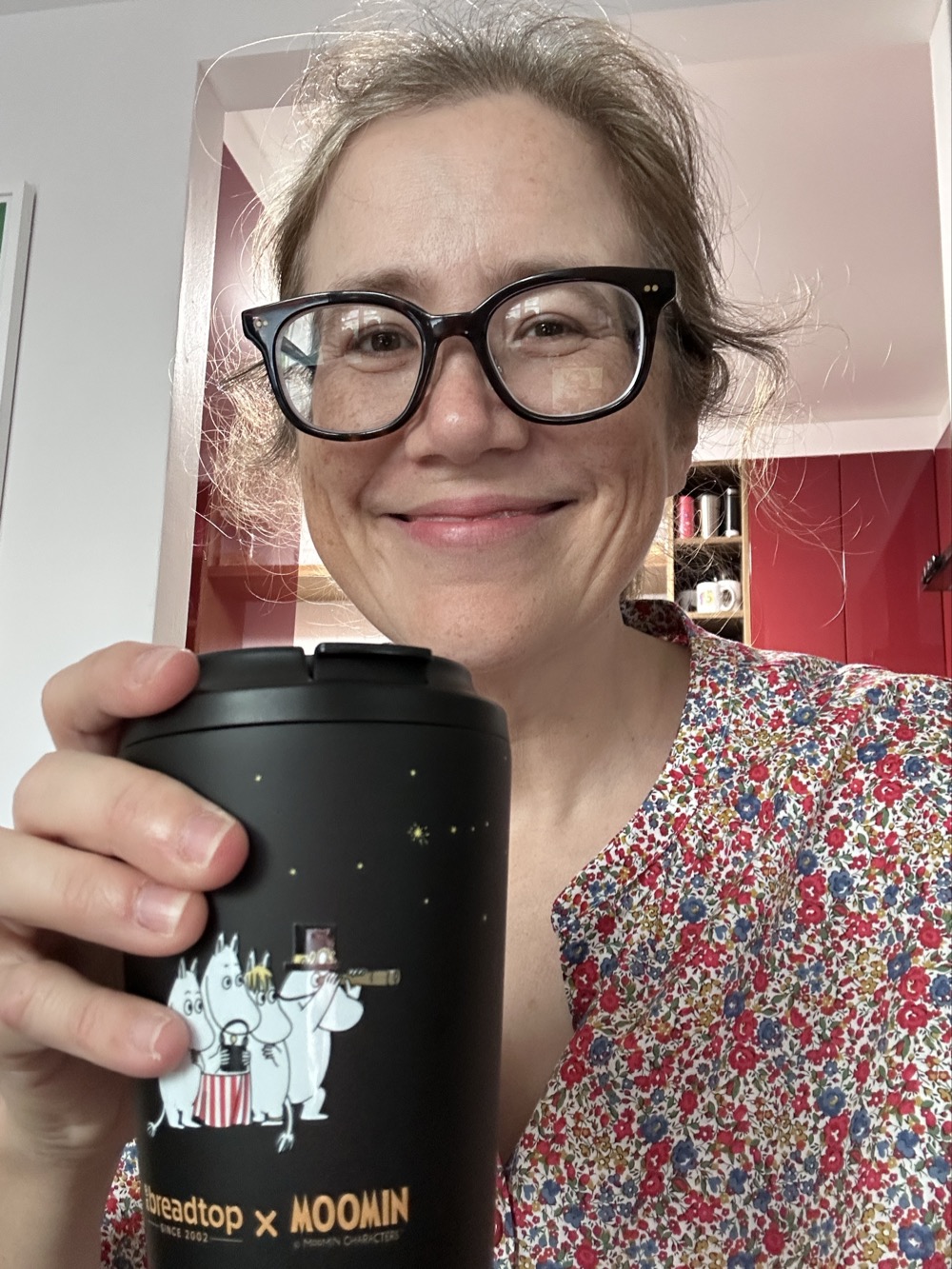 A woman in glasses smiling while holding a black reusable coffee cup with an illustration of Moomins on it